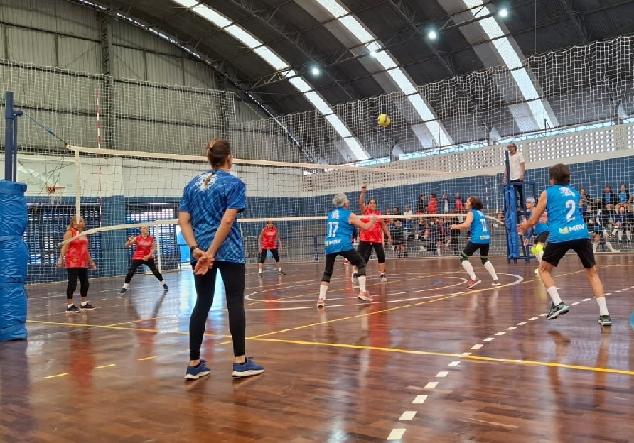 A imagem mostra uma partida de vôlei indoor em andamento, vista de um ângulo baixo e atrás de uma das equipes. O foco principal está na rede, onde a bola amarela está no ar, prestes a ser disputada. Os jogadores do time mais próximo usam uniformes vermelhos com shorts ou leggings pretas, enquanto os adversários vestem camisetas azuis com números brancos e o logo "MRV". No primeiro plano, uma mulher com camisa azul estampada e calça preta observa a partida de costas para a câmera, possivelmente uma técnica ou árbitra auxiliar. O ginásio tem piso de madeira polida com marcações brancas e amarelas, teto alto em formato de arco feito de metal ondulado e iluminação artificial forte. Ao fundo, há arquibancadas com alguns espectadores e um árbitro na plataforma ao lado da rede. Os jogadores parecem ser adultos de idade mais avançada, indicando que pode ser uma liga recreativa ou para a terceira idade. A cena transmite a energia e o movimento típicos de um jogo competitivo e amistoso de vôlei.