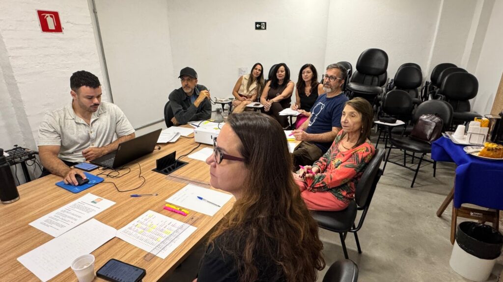 A imagem mostra um grupo de oito pessoas reunidas em uma sala clara, provavelmente para uma reunião ou workshop. Elas estão sentadas ao redor de uma mesa grande de madeira, que está coberta com papéis, canetas, marcadores e um celular. À esquerda, um homem com camisa polo branca está usando um laptop e um mouse. Em primeiro plano, uma mulher com cabelos longos e óculos é vista de perfil, olhando para o centro da mesa. Do outro lado da mesa, há seis pessoas sentadas, incluindo homens e mulheres de diferentes idades, todos atentos e focados na atividade. A sala tem paredes brancas e iluminação artificial forte. Ao fundo, há várias cadeiras pretas empilhadas e uma mesa coberta com um pano azul que tem alguns lanches e bebidas. Na parede, é possível ver uma placa de extintor de incêndio e uma placa de saída de emergência. O ambiente transmite uma atmosfera profissional e colaborativa.