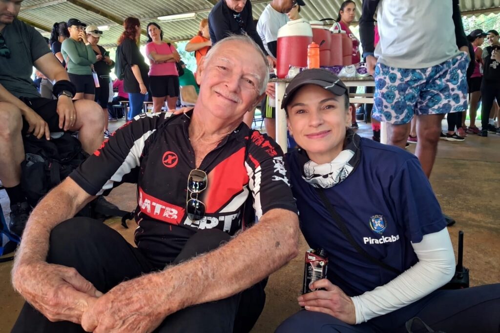 A imagem mostra um homem idoso e uma mulher de meia-idade sentados juntos no chão, sorrindo para a câmera. O homem está vestindo uma camisa de ciclismo nas cores preta, vermelha e branca, com a sigla "MTB" visível na frente, e usa óculos escuros presos na gola da camisa. A mulher usa uma camiseta azul escura com o brasão e a palavra "Piracicaba", além de mangas protetoras brancas nos braços e um boné cinza. Eles parecem estar participando de um evento ao ar livre, possivelmente relacionado a ciclismo ou atividade esportiva, sob uma estrutura coberta. Ao fundo, há várias pessoas vestindo roupas esportivas, algumas próximas a uma mesa com dispensadores de água vermelhos. A iluminação é natural e intensa, indicando que é durante o dia. A atmosfera da imagem é alegre e comunitária.