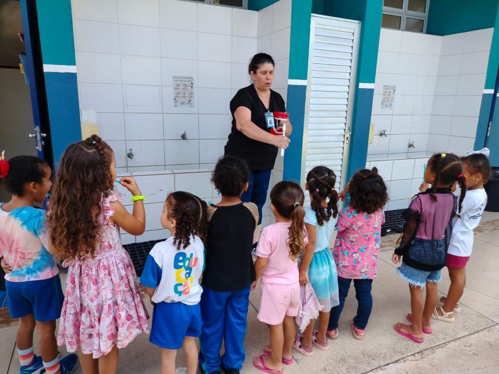 A imagem mostra uma cena educativa ao ar livre, onde uma mulher adulta, provavelmente uma professora ou profissional de saúde, está demonstrando técnicas de higiene bucal para um grupo de nove crianças pequenas. Ela segura um modelo grande de dentes e uma escova de dentes para explicar como escovar corretamente. As crianças, de diversas etnias, estão em semicírculo, prestando atenção à explicação. Elas vestem roupas coloridas e casuais, e uma delas usa uma camiseta com a palavra "Educação" estampada. O ambiente parece ser o pátio de uma escola, com paredes azuis e azulejos brancos, além de várias pias e torneiras ao fundo. Há também cartazes educativos sobre lavar as mãos, indicando um foco em saúde e higiene. A cena transmite um momento de aprendizado prático e interação entre educadora e alunos.