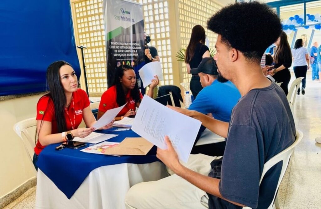 A imagem mostra uma cena típica de uma feira de empregos ou um evento de recrutamento, realizada em um ambiente interno, possivelmente um centro comunitário ou uma instituição pública. No centro da imagem, um jovem com cabelo afro volumoso está sentado à mesa, segurando um documento branco, aparentemente preenchendo ou revisando um formulário. À sua frente, duas mulheres vestindo camisas polo vermelhas com um distintivo da bandeira do Brasil e crachás, parecem ser recrutadoras ou representantes da empresa, analisando documentos e conversando com o candidato. Na mesa, há diversos papéis, pastas e um celular, indicando que estão realizando algum tipo de triagem ou registro. Ao fundo, um banner da empresa "SMS Scrap Metal Services" destaca que se trata de uma multinacional líder no setor de reciclagem de sucata metálica e serviços de siderurgia, sugerindo que a vaga oferecida está relacionada a essa área. O ambiente é bem iluminado, com uma parede decorativa ao fundo e outras pessoas circulando ou aguardando atendimento, o que reforça a ideia de um evento com vários candidatos. A imagem transmite a seriedade e o profissionalismo do processo seletivo, além do interesse dos jovens em oportunidades de trabalho.