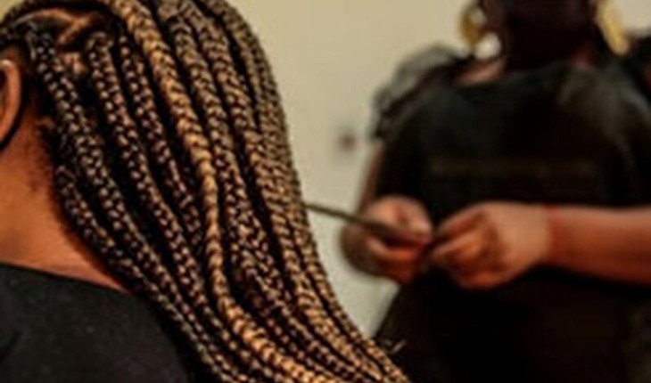 A imagem mostra uma pessoa vista de perfil, com o foco principal em seu cabelo trançado em box braids. As tranças são longas, bem feitas e uniformes, destacando-se em tons de preto e marrom. Ao fundo, desfocado, há outra pessoa, possivelmente um cabeleireiro, segurando uma ferramenta fina, provavelmente para ajudar na finalização das tranças. O ambiente parece ser interno, talvez um salão de beleza ou uma área de cuidados capilares em casa. A iluminação é suave, realçando a textura das tranças, enquanto o fundo está desfocado para destacar o cabelo da pessoa em primeiro plano.