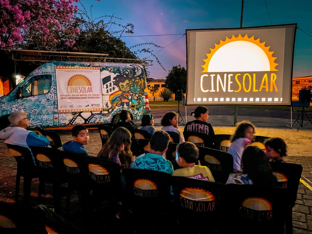 A imagem mostra um evento ao ar livre chamado "CINESOLAR", que é uma estação solar móvel de cinema. Vemos várias pessoas, principalmente crianças, sentadas em cadeiras pretas com o logotipo do CINESOLAR, assistindo a um filme projetado em uma grande tela branca. O evento ocorre durante o entardecer, com o céu em tons de azul e roxo, criando uma atmosfera tranquila. À esquerda da imagem, há uma van decorada com arte colorida e um banner que diz "CINESOLAR ESTAÇÃO SOLAR MÓVEL DE CINEMA ARTE.SUSTENTABILIDADE.TECNOLOGIA". Essa van provavelmente funciona como a estrutura móvel que alimenta o cinema com energia solar, promovendo sustentabilidade. Ao fundo, é possível ver árvores e algumas construções, sugerindo que o evento ocorre em uma área aberta, possivelmente um parque ou praça. A iluminação principal vem da tela do cinema e da van, destacando os espectadores e o ambiente ao redor. O evento parece ser uma iniciativa do Governo do Estado de São Paulo, conforme indicado no banner. A cena transmite uma sensação de comunidade, cultura e tecnologia sustentável.