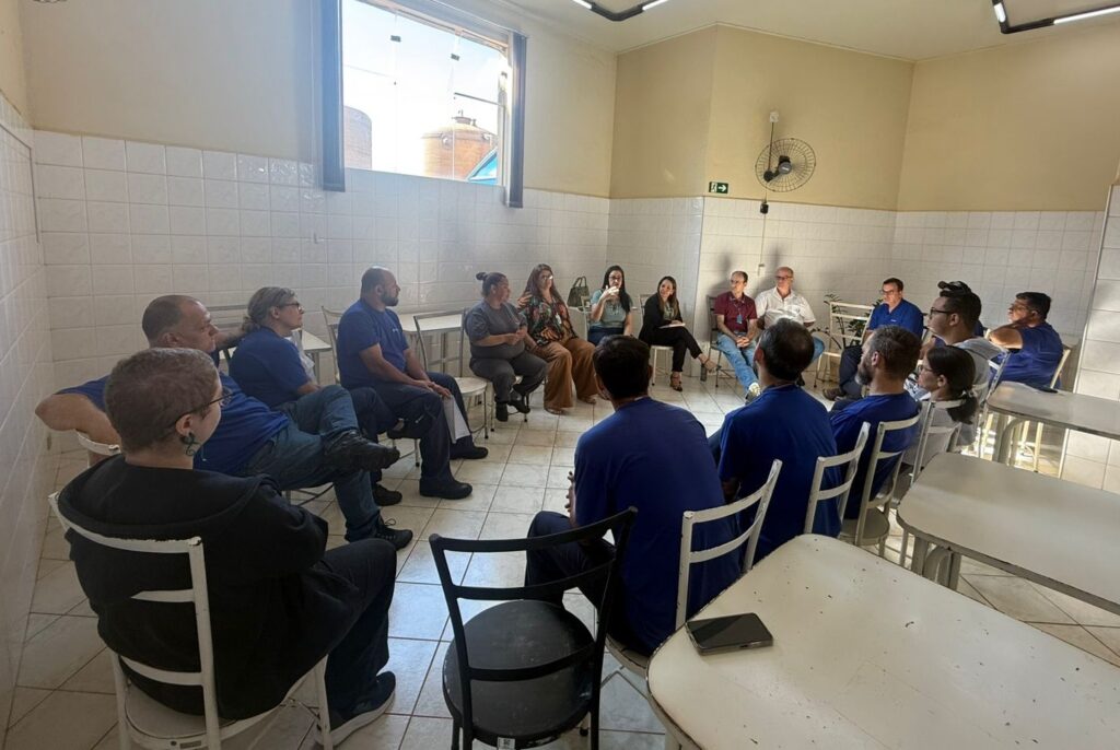 A imagem mostra um grupo de aproximadamente 16 pessoas sentadas em círculo dentro de uma sala que parece ser uma cantina ou sala de descanso de uma empresa. A maioria dos homens está vestindo camisetas azuis, possivelmente uniformes, enquanto as mulheres usam roupas casuais ou profissionais variadas. Eles parecem estar participando de uma reunião ou discussão, com algumas pessoas prestando atenção a um interlocutor. A sala é clara, com paredes brancas e piso de azulejos claros. Há janelas grandes que deixam entrar bastante luz natural, e uma parede possui um ventilador fixado. As cadeiras são brancas, de metal, com encosto vazado, e as mesas foram empurradas para as laterais da sala para dar espaço ao círculo central. Do lado de fora da janela, é possível ver estruturas industriais, como silos, indicando que o local pode ser uma área industrial ou de fábrica. No geral, a imagem transmite a ideia de uma reunião de equipe, talvez uma sessão de treinamento, briefing de segurança ou discussão de trabalho em um ambiente industrial.