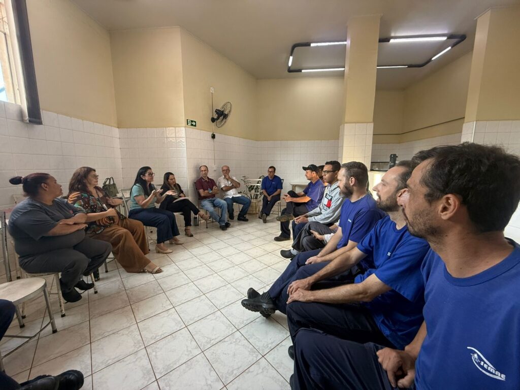 A imagem mostra um grupo de pessoas reunidas em uma sala, sentadas em círculo, aparentemente participando de uma reunião ou discussão. De um lado, há várias mulheres, e do outro, homens vestidos com uniformes azuis que têm a sigla "SEMAE" nas costas. A sala tem paredes com azulejos brancos na parte inferior e pintura bege na parte superior. O ambiente parece ser simples, com cadeiras comuns e iluminação fluorescente no teto. Também é possível ver um ventilador de parede e uma placa de saída no fundo. A disposição das pessoas sugere um momento de diálogo ou treinamento, com atenção voltada para quem está falando. O clima é profissional, mas informal.