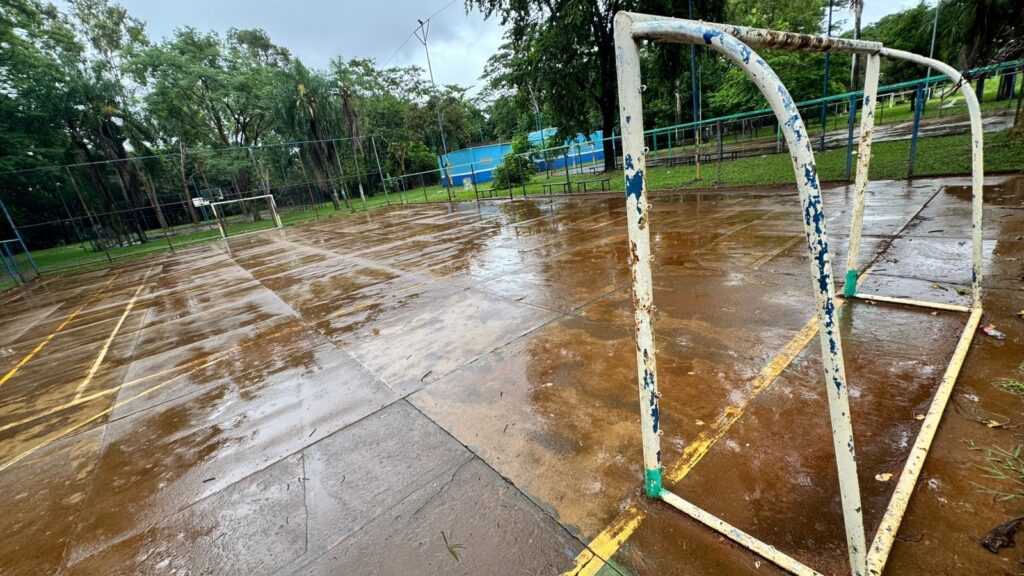 A imagem mostra uma quadra esportiva ao ar livre, provavelmente usada para futebol de salão ou basquete, em um dia chuvoso. O chão da quadra está molhado, com poças de água refletindo a luz do céu nublado. Em primeiro plano, há um gol de metal branco com pintura azul e verde desgastada, apresentando ferrugem. Ao fundo, há outro gol menor, também branco. A quadra é cercada por uma cerca de arame verde, e ao redor há muitas árvores verdes, sugerindo um ambiente de parque ou área recreativa. O céu está cinza e nublado, indicando que a chuva caiu recentemente ou ainda está caindo. Não há pessoas na cena, e a atmosfera transmite uma sensação de tranquilidade e abandono momentâneo devido ao mau tempo.