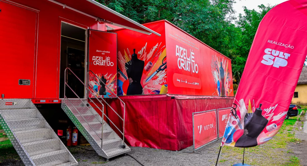 A imagem mostra uma unidade móvel grande e vibrante, provavelmente um trailer adaptado para funcionar como um ateliê criativo itinerante. O trailer é predominantemente vermelho e possui diversas ilustrações relacionadas ao universo da costura, como tesouras, bobinas de linha e manequins, destacando seu propósito artístico e artesanal. Na lateral do trailer, há uma escada metálica com corrimão que leva até uma porta aberta, sugerindo que o espaço está pronto para receber visitantes ou alunos. Ao lado do trailer, há uma bandeira vertical rosa com inscrições que remetem à organização responsável pelo projeto, "REALIZAÇÃO CULT SP", e elementos gráficos semelhantes aos do trailer. O cenário é ao ar livre, com árvores verdes ao fundo e um céu nublado, indicando um dia sem sol forte. No chão, há uma mistura de terra e cascalho, e o trailer está posicionado próximo a uma via asfaltada. Pequenas placas vermelhas com a palavra "VETOR" também estão visíveis próximas ao trailer. O texto na lateral do trailer diz: "Bem-vindos ao ATELIÊ CRIATIVO. Governo do Estado de São Paulo por meio da Secretaria de Cultura, Economia e Indústria Criativas apresenta: ATELIÊ CRIATIVO." Isso indica que o projeto é uma iniciativa oficial do governo para promover atividades culturais e criativas. Em resumo, a imagem retrata um espaço móvel dedicado à criatividade e à arte, especialmente relacionado à costura e moda, com o objetivo de levar essas atividades para diferentes localidades.