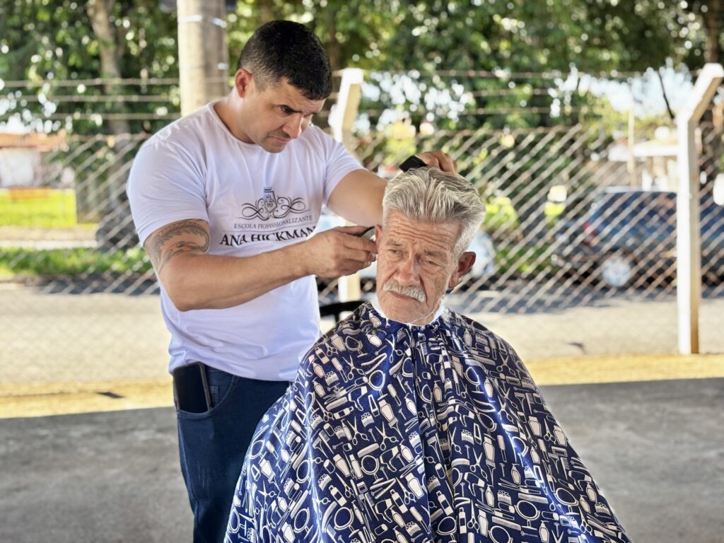 A imagem mostra um barbeiro cortando o cabelo de um homem idoso ao ar livre. O barbeiro é um homem de meia-idade, com cabelo curto e escuro, usando uma camiseta branca com um logo e calça azul escura. Ele está concentrado no corte, segurando um pente na mão esquerda e uma máquina de cortar cabelo na mão direita. O homem idoso tem cabelos grisalhos, ondulados, e bigode branco, vestindo uma capa azul com desenhos de tesouras, pentes e outros instrumentos de barbearia. Ao fundo, há uma cerca de arame e árvores verdes, indicando que o corte está acontecendo em um ambiente externo, possivelmente um parque ou praça. A iluminação é natural e clara, sugerindo que é durante o dia. No bolso traseiro do barbeiro, há um celular preto. Na imagem também aparece a inscrição "ESCOLA PROFISSIONALIZANTE ANA HICKMANN", indicando que o local pode estar relacionado a uma escola de formação profissional.