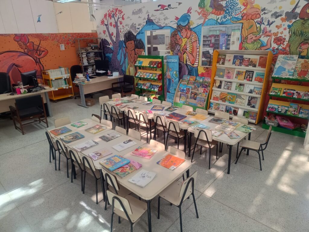 A imagem retrata uma biblioteca ou sala de leitura infantil, cheia de cores e elementos lúdicos. No centro, várias mesas pequenas estão agrupadas, cercadas por cadeirinhas, e cobertas com livros e revistas de capas ilustradas e vibrantes. Ao fundo, prateleiras bem organizadas exibem uma grande coleção de livros infantis. As paredes são decoradas com murais grandes e coloridos, trazendo personagens em estilo de desenho animado, cenas da natureza e elementos imaginativos que tornam o ambiente acolhedor e estimulante. À esquerda, há mesas com computadores, papéis e materiais de escritório, sugerindo um espaço de apoio para bibliotecários ou funcionários.