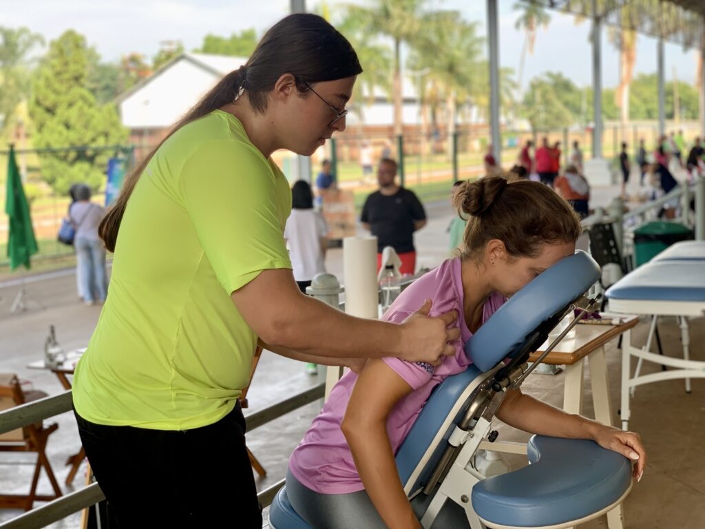 A imagem mostra uma terapeuta de massagem, vestindo uma camiseta verde-limão, realizando uma massagem em cadeira em uma cliente que está sentada de frente para a cadeira, com o rosto apoiado no apoio acolchoado. A cliente usa uma blusa roxa e tem cabelo preso em um coque. A terapeuta está posicionada atrás e um pouco ao lado da cliente, aplicando a massagem nos ombros dela. O ambiente parece ser ao ar livre, sob uma cobertura em uma área que lembra um espaço esportivo ou uma feira, com luz natural e árvores ao fundo. Há outras pessoas e cadeiras de massagem desfocadas ao fundo, sugerindo um evento comunitário de saúde e bem-estar. A atmosfera é tranquila e profissional.