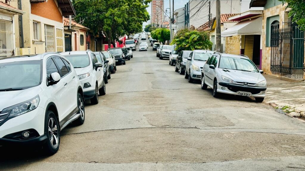 A imagem mostra uma rua residencial estreita e inclinada, ladeada por carros estacionados dos dois lados. No primeiro plano, à esquerda, há um SUV branco da marca Honda, e à direita, um carro prata, um Peugeot, com a placa "EAW-5353" visível. A rua se estende em direção ao fundo da imagem, onde é possível ver mais veículos estacionados e algumas árvores. As casas são de tijolos aparentes e há postes de energia elétrica com fios passando pelo alto da rua. A cena parece ser em um dia claro, porém com céu nublado, e não há pessoas visíveis na imagem. O ambiente transmite uma sensação típica de um bairro urbano tranquilo.