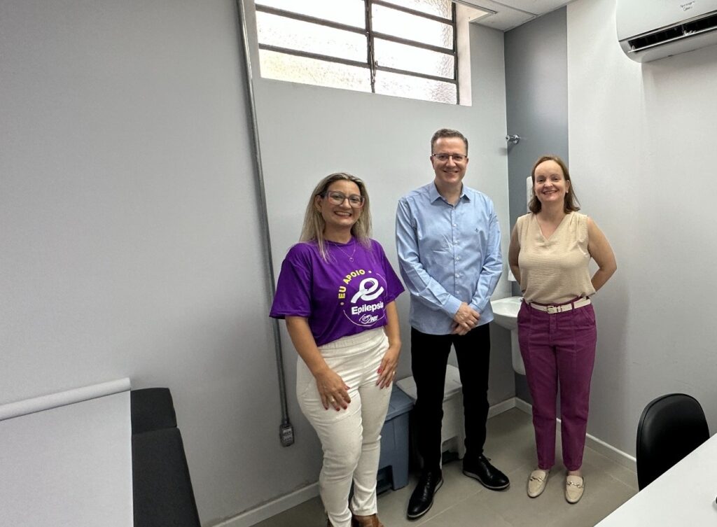 A imagem mostra três pessoas em um ambiente que parece ser um consultório médico ou clínico. À esquerda, há uma mulher loira usando óculos, camiseta roxa com um logotipo e calça branca. No centro, um homem com cabelo castanho curto e óculos veste uma camisa azul clara e calça preta. À direita, uma mulher com cabelo castanho claro usa uma blusa bege sem mangas e calça roxa. Todos estão sorrindo e olhando para a câmera, posando para a foto. O ambiente é simples, com paredes cinza, uma janela pequena alta com vidro fosco e um aparelho de ar-condicionado no canto superior direito. À esquerda, há uma mesa de exame branca, e ao fundo, pode-se ver uma pia e lixeiras. A camiseta roxa da mulher à esquerda tem um logotipo com a inscrição "EU APOIO Epilepsia", sugerindo que o local ou o grupo está relacionado ao apoio a pessoas com epilepsia. A iluminação é clara e típica de ambientes internos.