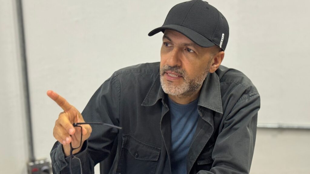 A imagem mostra um homem de meia-idade, com pele morena e barba grisalha, aparentando estar entre 40 e 50 anos. Ele veste um boné preto com a palavra "KIPSTA" escrita em branco na lateral, uma camisa escura por cima de uma camiseta azul. O homem está olhando para a esquerda, com uma expressão pensativa e atenta, como se estivesse falando ou explicando algo. Sua mão direita está levantada, com o dedo indicador apontando para cima, segurando também um par de óculos de armação preta. O fundo é simples, com uma parede clara e alguns elementos metálicos, sugerindo um ambiente interno, possivelmente um escritório ou sala de aula. A iluminação é suave, destacando os detalhes do rosto e da roupa. A composição transmite uma sensação de diálogo ou reflexão.