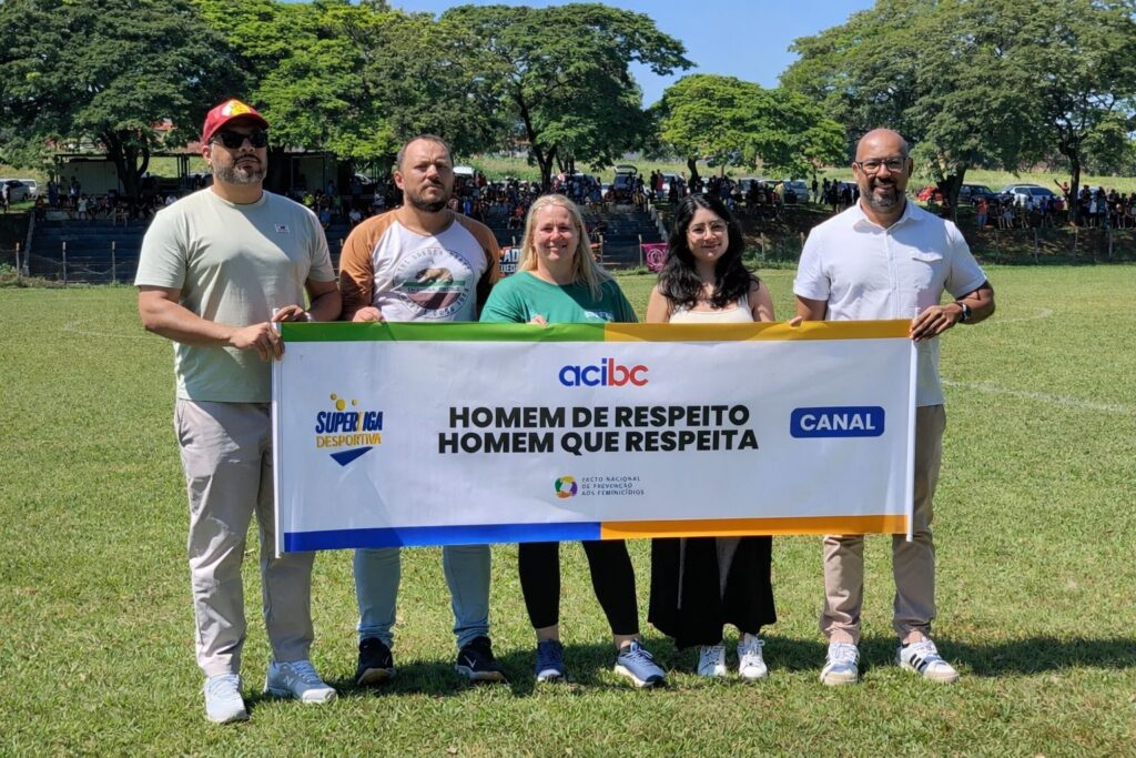 A imagem mostra cinco adultos em pé, alinhados lado a lado em um campo de grama sob a luz do sol. Eles seguram uma faixa branca com bordas coloridas e o texto em destaque: "HOMEM DE RESPEITO HOMEM QUE RESPEITA". Na faixa também aparecem os logos de várias organizações, incluindo "acibc", "SUPERLIGA DESPORTIVA", "CANAL" e "PACTO NACIONAL DE PREVENÇÃO AOS FEMINICÍDIOS". As pessoas parecem estar participando de um evento ao ar livre, possivelmente relacionado a esportes ou uma campanha social, dada a presença da multidão ao fundo, sentada em arquibancadas. O cenário ao redor é verde, com árvores grandes e um céu claro, indicando um dia ensolarado. A mensagem da faixa sugere um foco na conscientização sobre respeito e prevenção da violência contra as mulheres.
