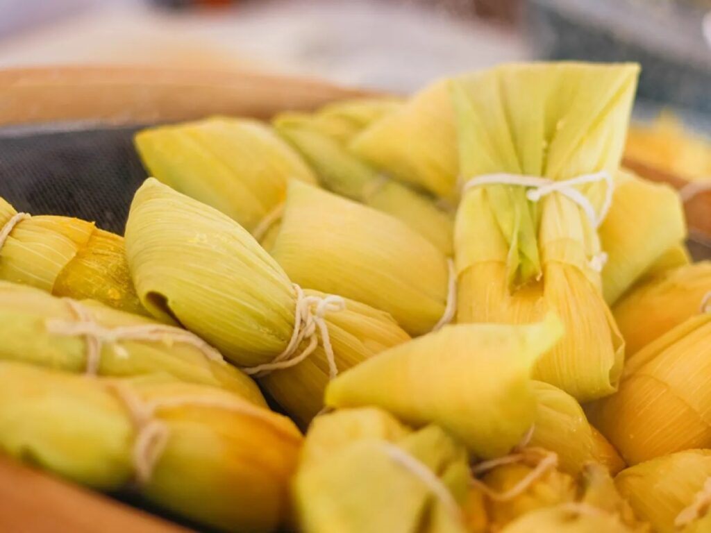 A imagem mostra um conjunto de tamales ou humitas envoltos em folhas de milho, amarrados com fios brancos. As folhas têm tons que variam do amarelo claro ao verde suave, exibindo texturas naturais e sulcos típicos do milho. Os tamales estão agrupados de forma compacta, preenchendo toda a imagem, com uma iluminação suave que destaca os detalhes das folhas. O fundo está desfocado, sugerindo um ambiente rústico, possivelmente uma cozinha ou local tradicional de preparo. A composição transmite uma sensação de abundância e autenticidade culinária.