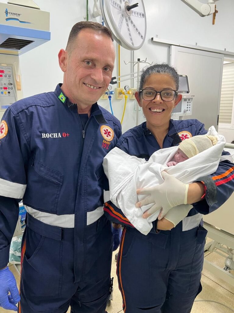A imagem mostra dois paramédicos em um ambiente hospitalar, provavelmente uma sala de parto ou emergência obstétrica. Ambos vestem uniformes azuis escuros com faixas refletivas prateadas. O paramédico à esquerda é um homem, com cabelo curto, e tem o nome "ROCHA" e o tipo sanguíneo "O+" bordados no uniforme, além do logo "SAMU 192" e uma pequena bandeira do Brasil na gola. A paramédica à direita é uma mulher, que usa óculos e luvas brancas, segurando um recém-nascido envolto em um cobertor branco e usando um gorro de tricô claro. Ao fundo, há equipamentos médicos, como um monitor, luz cirúrgica e tomadas de gases medicinais, caracterizando um ambiente clínico. A expressão dos paramédicos é de alegria e satisfação, sugerindo que o nascimento do bebê ocorreu com sucesso e eles estão comemorando o momento.