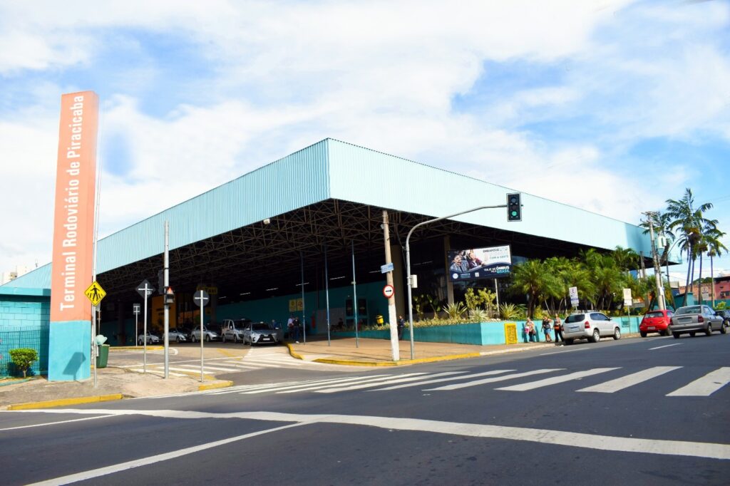 A imagem mostra o Terminal Rodoviário de Piracicaba, um grande terminal de ônibus com um design moderno e industrial. O destaque é o amplo telhado em balanço, que cobre a área de embarque, com uma estrutura metálica visível. À esquerda, há um totem alto e colorido nas cores laranja e azul, com o nome do terminal escrito. Na frente do terminal, há uma via asfaltada com algumas faixas de pedestres e sinais de trânsito. Vários carros estão estacionados ou circulando pela área, incluindo um táxi, indicado por placas específicas. O céu está claro, com poucas nuvens, e algumas palmeiras aparecem do lado direito, sugerindo um ambiente urbano tropical. Pessoas pequenas podem ser vistas caminhando perto da entrada do terminal. O ambiente transmite a ideia de um local organizado e funcional, destinado ao transporte rodoviário de passageiros.