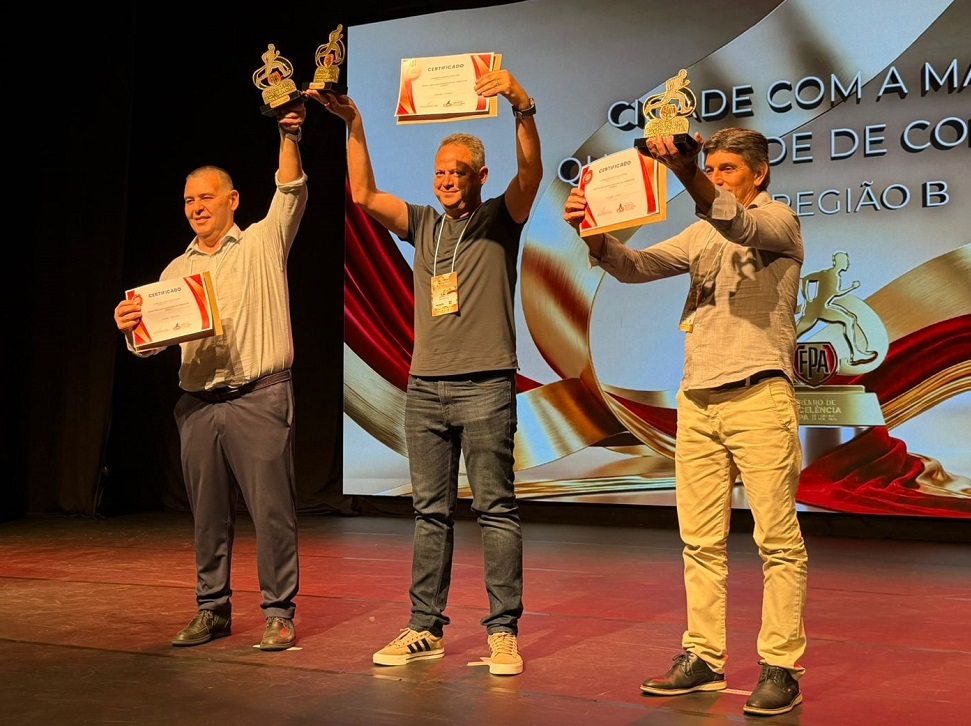 A imagem mostra três homens em um palco de madeira escura, durante uma cerimônia de premiação. Eles estão alinhados, sorrindo e segurando troféus dourados e certificados. O homem à esquerda veste uma camisa clara e calças escuras, segurando dois troféus e um certificado. O homem do meio usa uma camiseta cinza e jeans, levantando seu certificado com ambas as mãos. O homem à direita veste uma camisa clara e calças cáqui, segurando um troféu e um certificado. Ao fundo, há uma tela grande com um design em tons dourados e vermelhos, exibindo o texto "CERTIFICADO", "CIDADE COM A MA", "QUALIDADE DE CO", "REGIÃO B", "FPA" e "PRÊMIO DE EXCELÊNCIA", indicando que eles receberam um prêmio de excelência, provavelmente relacionado a atletismo, dado o logo da Federação Paulista de Atletismo (FPA) e os troféus com figuras de corredores. A iluminação destaca os premiados, enquanto o restante do ambiente está mais escuro, típico de eventos em auditórios ou teatros. A atmosfera parece de celebração e reconhecimento.