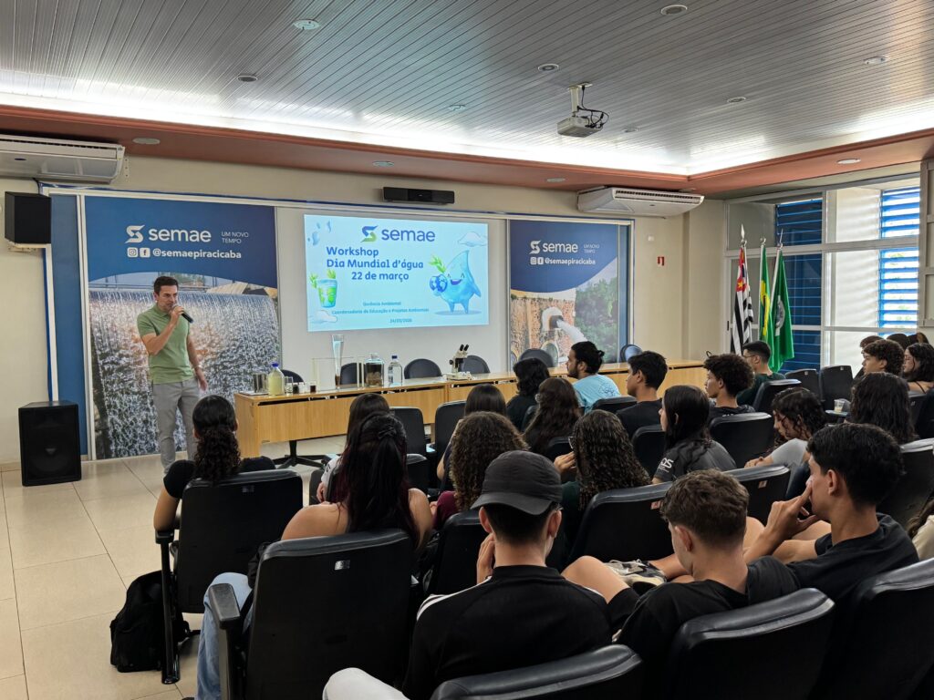Esta imagem mostra um ambiente interno, provavelmente uma sala de conferências ou auditório, onde está acontecendo um workshop sobre o Dia Mundial da Água, celebrado em 22 de março. Um homem, que parece ser o palestrante, está em pé à esquerda da imagem, segurando um microfone e falando para o público. Ele usa uma camisa polo verde claro e calça cinza. À sua frente, há um telão grande exibindo a apresentação do evento, que tem o título "Workshop Dia Mundial d'água 22 de março" e o logo do "semae". Na tela também aparece um desenho de uma gota d’água com olhos e boca, dando um aspecto lúdico à apresentação. O público é formado por cerca de 20 a 25 pessoas, principalmente jovens adultos, que estão sentados em cadeiras pretas e prestam atenção ao palestrante. Atrás deles, à direita, há janelas com persianas azuis e três bandeiras, uma delas provavelmente do Brasil. Na frente do palco, sobre uma mesa comprida de madeira, estão expostos diversos objetos relacionados ao tema da água, como frascos com líquidos, um microscópio e outros materiais didáticos. As paredes têm banners com o logo do semae e imagens relacionadas à água e saneamento. O ambiente está bem iluminado com luz branca e o teto é revestido com placas brancas e luminárias embutidas. No geral, a cena retrata uma atividade educativa e informativa sobre a importância da água, promovida por uma organização ligada ao saneamento ambiental.