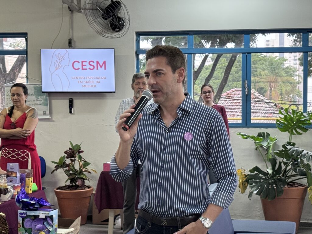 A imagem mostra um homem de meia-idade falando ao microfone, provavelmente durante um evento ou apresentação. Ele veste uma camisa azul com listras brancas e usa um relógio prateado no pulso esquerdo. Atrás dele, há uma tela digital azul com o texto "CESM CENTRO ESPECIALIZA EM SAÚDE DA MULHER", acompanhada de um logo estilizado de perfil feminino. Ao lado esquerdo do homem, uma mulher de vestido vermelho observa a cena com os braços cruzados. Ao fundo, em uma sala bem iluminada e decorada com plantas (como uma costela-de-adão e uma planta com folhas coloridas), aparecem outras duas pessoas, um homem e uma mulher, ambos com expressão atenta. A janela grande com moldura azul deixa entrar luz natural e mostra árvores e telhados ao fundo, indicando que o local é um ambiente interno, possivelmente um centro de saúde ou uma clínica especializada em atendimento à mulher. O clima da imagem sugere um momento formal, possivelmente uma palestra, anúncio ou cerimônia relacionada à saúde feminina.