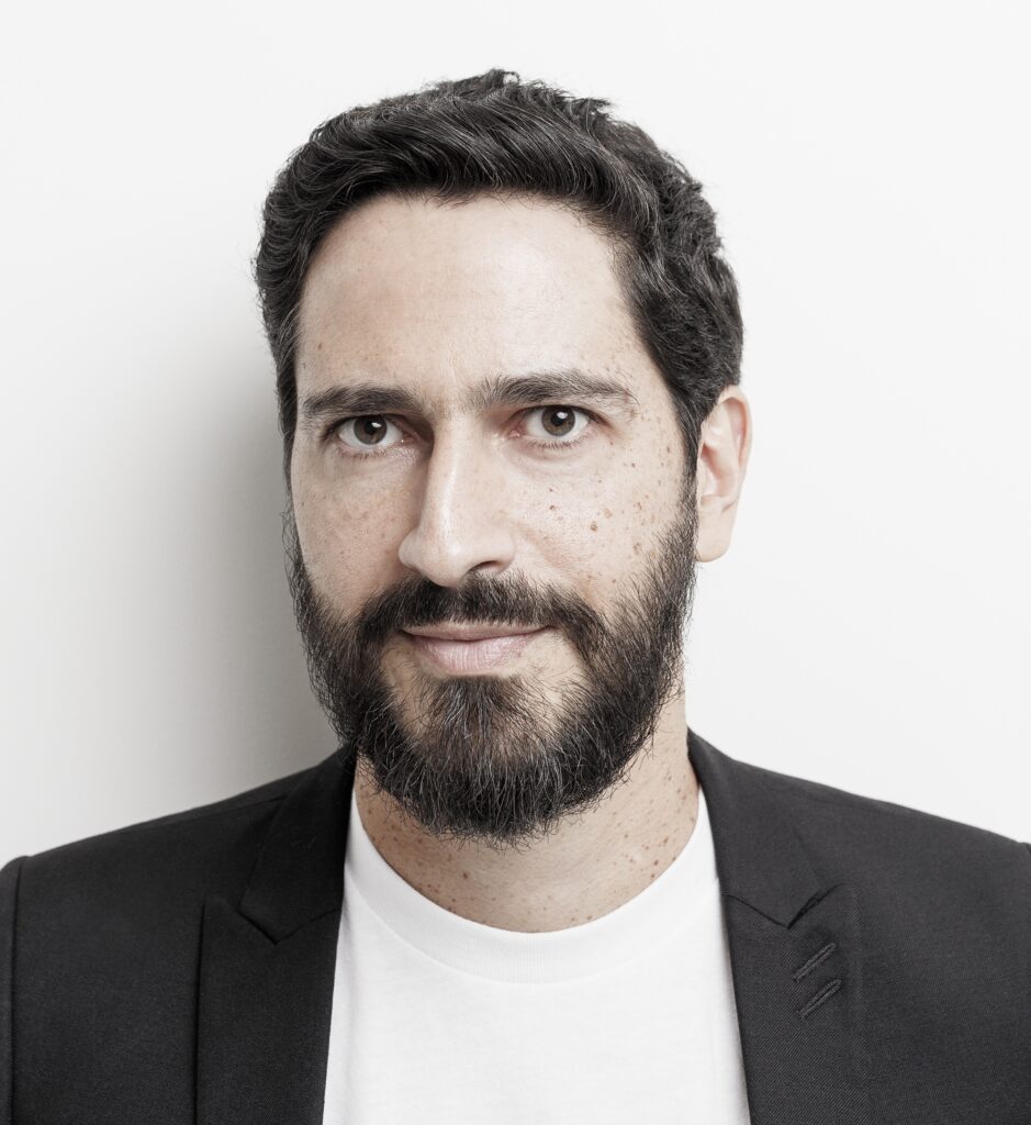 A imagem mostra o retrato de um homem de meia-idade, visto da altura dos ombros para cima. Ele tem cabelos escuros, levemente ondulados, e uma barba cheia e bem cuidada, também escura. Seu rosto apresenta algumas sardas visíveis nas bochechas e na testa. O homem está vestido de forma casual, usando uma camiseta branca básica por baixo de um blazer preto. O fundo é completamente branco, o que destaca ainda mais o rosto e a figura dele. A iluminação é clara e uniforme, sem sombras fortes, sugerindo que a foto foi tirada em estúdio. A expressão dele é neutra, com um leve sorriso, transmitindo uma sensação de simpatia e profissionalismo.