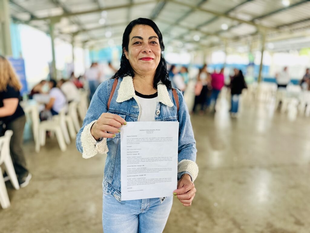 A imagem mostra uma mulher sorridente, de cabelos escuros, vestindo uma jaqueta jeans com forro branco e uma camiseta branca por baixo, segurando um currículo impresso. Ela está em um ambiente amplo e coberto, com estrutura metálica e várias pessoas ao fundo, sugerindo um evento como uma feira de emprego ou reunião comunitária. O currículo que ela segura pertence a Rosimara Cristina Gonçalves, de 49 anos, residente em Piracicaba, SP. No currículo, Rosimara apresenta seu objetivo profissional, destacando sua experiência como camareira, profissional de serviços gerais e cuidadora de idosos, enfatizando seu compromisso com a qualidade e o bem-estar dos clientes. Ela possui ensino médio completo desde 2000. Sua experiência inclui trabalhos como camareira em um motel, auxiliar de produção em curtume e cuidadora de idosos, com responsabilidades detalhadas em cada função. Também menciona disponibilidade para início imediato. A imagem e o currículo transmitem uma impressão de profissional dedicada e pronta para novas oportunidades de trabalho.