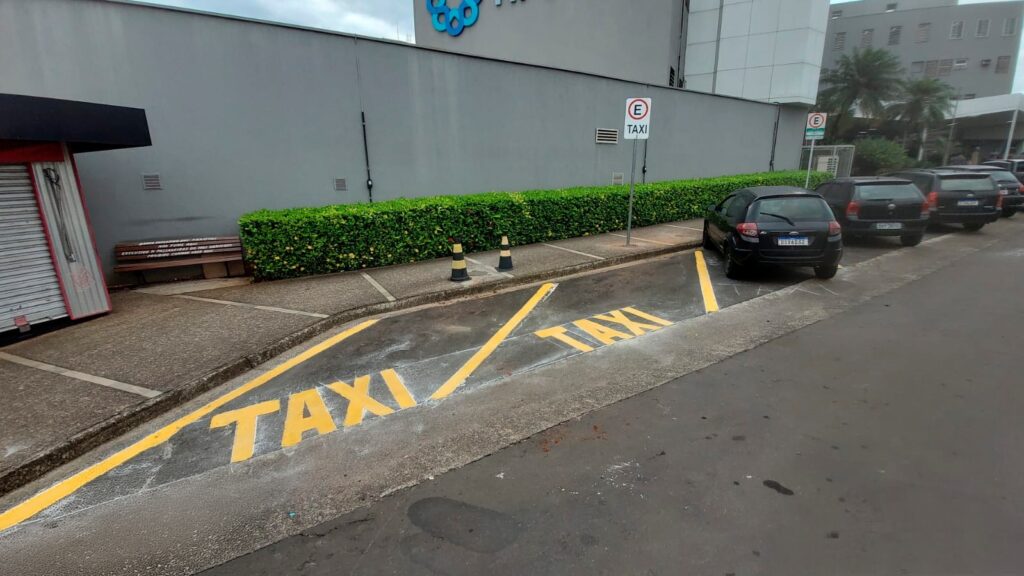 A imagem mostra uma área de estacionamento exclusiva para táxis em frente a um prédio moderno. No chão, há marcações amarelas indicando a vaga para táxi, com a palavra "TAXI" escrita em destaque. Um carro preto, modelo Ford Ka, está estacionado na vaga, com a placa DSX6D32. Ao lado, há outros veículos estacionados, incluindo um Volkswagen preto. Na calçada, há um banco de madeira com uma placa que contém várias informações em português, como "BANCO EXCLUSIVO PARA TAXISTAS", "NÃO FUME AQUI", "EXCLUSIVO P/ PASSAGEIROS DO TAXI AGRADE" e "PROIBIDO COMER NESTE LOCAL", indicando regras específicas para o uso do espaço. Também há cones de trânsito laranja e preto posicionados perto do banco. Ao fundo, vê-se um quiosque fechado com uma porta metálica branca e vermelha, e um muro com vegetação baixa ao longo da calçada. Placas indicativas com a letra "E" dentro de um círculo e a palavra "TAXI" reforçam que a área é destinada exclusivamente para táxis. A cena aparenta ser em um ambiente urbano, provavelmente no Brasil, devido às placas e idioma utilizados.