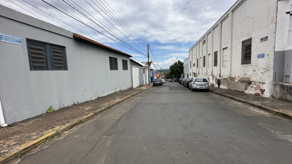 A imagem mostra uma rua estreita e pavimentada, vista em perspectiva, que se estende em direção a um ponto de fuga no horizonte. De ambos os lados da rua, há prédios baixos, com fachadas simples e algumas janelas, que parecem ser de uso residencial ou comercial. A iluminação é clara, indicando que a foto foi tirada durante o dia, provavelmente em um dia nublado ou com luz difusa. No lado direito da imagem, há vários carros estacionados ao longo da calçada, incluindo um carro prata da marca Renault em destaque próximo ao primeiro plano. No lado esquerdo, há um carro escuro estacionado mais distante. Acima da rua, fios elétricos cruzam o céu, que está parcialmente visível e apresenta um tom claro. Há placas de rua visíveis em dois pontos: uma na parede do lado esquerdo e outra no lado direito, ambas com o nome "Nossa Senhora de Lourdes", indicando o nome da rua. A placa do lado esquerdo é azul, enquanto a do lado direito é preta com letras brancas. A cena transmite a sensação de um bairro urbano ou industrial tranquilo em uma cidade brasileira, com uma atmosfera calma e sem movimento, já que não há pessoas visíveis na rua. A composição da foto, com a rua centralizada e os prédios alinhados, cria um efeito de profundidade que guia o olhar para o fundo da imagem.