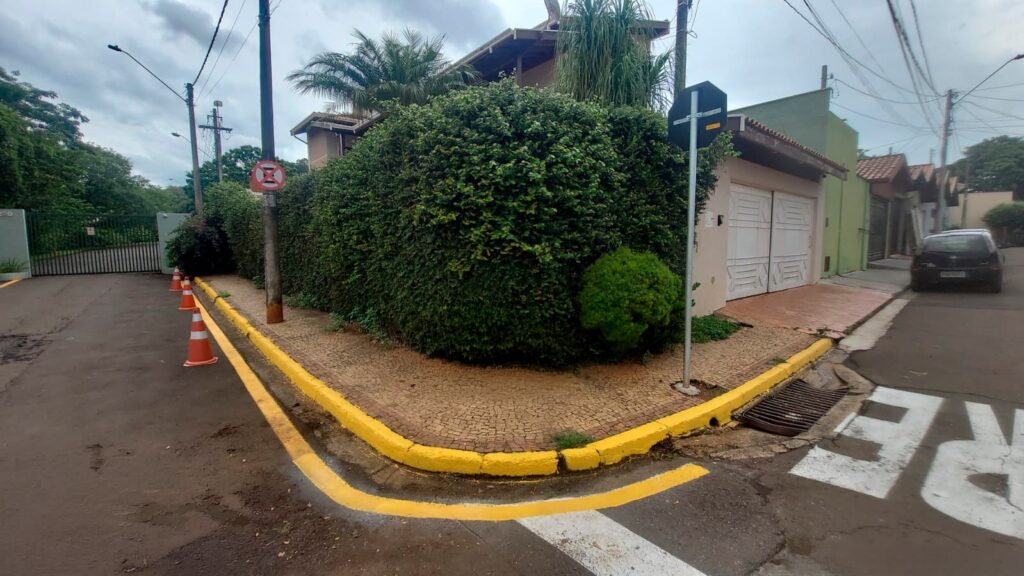 A imagem mostra uma esquina de rua em uma área residencial. No centro da cena, há uma grande cerca viva verde muito densa, bem cuidada, que ocupa boa parte do terreno. Em frente à cerca, o meio-fio está pintado de amarelo, indicando uma área de estacionamento proibido ou atenção especial. Na calçada, há alguns cones de trânsito laranja posicionados próximos a um portão branco, sugerindo que a entrada pode estar temporariamente bloqueada ou sinalizada. No chão da rua, está pintada a palavra "PARE" em letras grandes e brancas, indicando uma placa de pare para os veículos que chegam à esquina. Ao fundo, é possível ver uma casa branca com garagem e um carro preto estacionado. O céu está nublado, sem muita luz do sol, o que deixa a cena com uma iluminação difusa e sem sombras fortes. No geral, a imagem transmite um ambiente tranquilo e organizado, típico de um bairro residencial. A presença da cerca viva alta sugere privacidade para a residência.