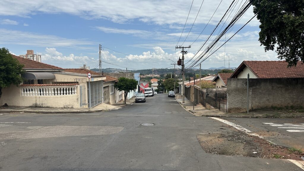 A imagem mostra uma rua residencial em uma área urbana com relevo inclinado. A rua de asfalto desce em direção ao horizonte, ladeada por casas de diferentes estilos. À esquerda, há uma casa clara com um portão branco e uma cerca com arame farpado no topo, além de uma árvore pequena. À direita, outras casas com telhados de cerâmica e paredes de concreto, parcialmente cobertas por uma grande árvore com folhagem densa. Vários carros estão estacionados ao longo da rua, incluindo um sedã prata e uma caminhonete branca mais ao fundo. Postes de energia com muitos fios atravessam a cena horizontalmente. O céu está azul com algumas nuvens brancas, indicando um dia ensolarado. No chão, próximo à parte inferior da imagem, há a palavra "PARE" pintada, que é um sinal de trânsito para os veículos. Ao fundo, é possível ver mais construções e colinas verdes distantes. A cena transmite uma sensação típica de um bairro tranquilo em uma cidade com relevo acidentado.