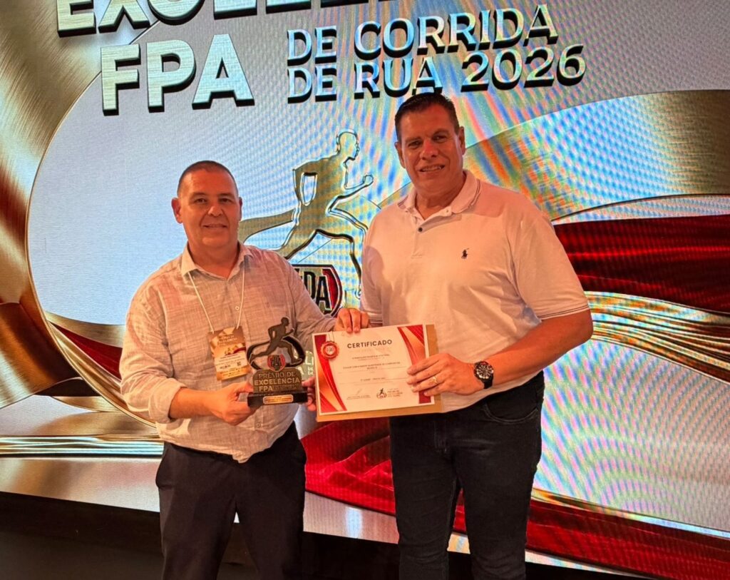 A imagem mostra uma cerimônia de premiação, onde dois homens estão posando para uma foto em frente a um grande painel digital. O painel exibe o texto "FPA DE CORRIDA DE RUA 2026" em letras grandes e metálicas, junto com a imagem estilizada de um corredor dourado e elementos gráficos em vermelho e dourado. O homem à esquerda veste uma camisa clara de manga curta e calça escura, segurando um troféu preto em formato de corredor. Ele também usa um crachá pendurado no pescoço. O homem à direita está vestido com uma camiseta polo branca e jeans escuros, segurando um certificado emoldurado que diz "CERTIFICADO" e indica que a cidade de Piracicaba conquistou o 2º lugar na categoria "Cidade com a maior quantidade de corridas na Região 4". Ambos sorriem para a câmera, transmitindo uma atmosfera de celebração e reconhecimento no contexto de corrida de rua. A iluminação é clara e uniforme, típica de eventos realizados em ambientes fechados.