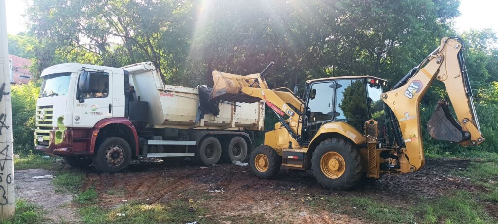 A imagem mostra uma cena de trabalho ao ar livre, com um caminhão basculante branco da marca Volkswagen Constellation, modelo FACCHINI, e uma retroescavadeira amarela da marca Caterpillar (CAT), modelo 416. A retroescavadeira está posicionada ao lado do caminhão, com a pá frontal levantada e despejando material escuro no compartimento de carga do caminhão. O caminhão tem a parte inferior da cabine pintada de vermelho escuro, e o compartimento de carga é branco. Ambos os veículos exibem o logotipo "ECOTERRA" com uma folha verde, indicando provavelmente a empresa responsável pelos equipamentos ou pela obra. O ambiente é um terreno de terra, com marcas de pneus e algumas sujeiras espalhadas pelo chão. Ao fundo, há árvores com folhagem densa, e a luz do sol cria um efeito de brilho, sugerindo que a foto foi tirada durante o dia, em um momento ensolarado. O cenário transmite uma sensação de trabalho pesado em uma área possivelmente rural ou em um canteiro de obras.