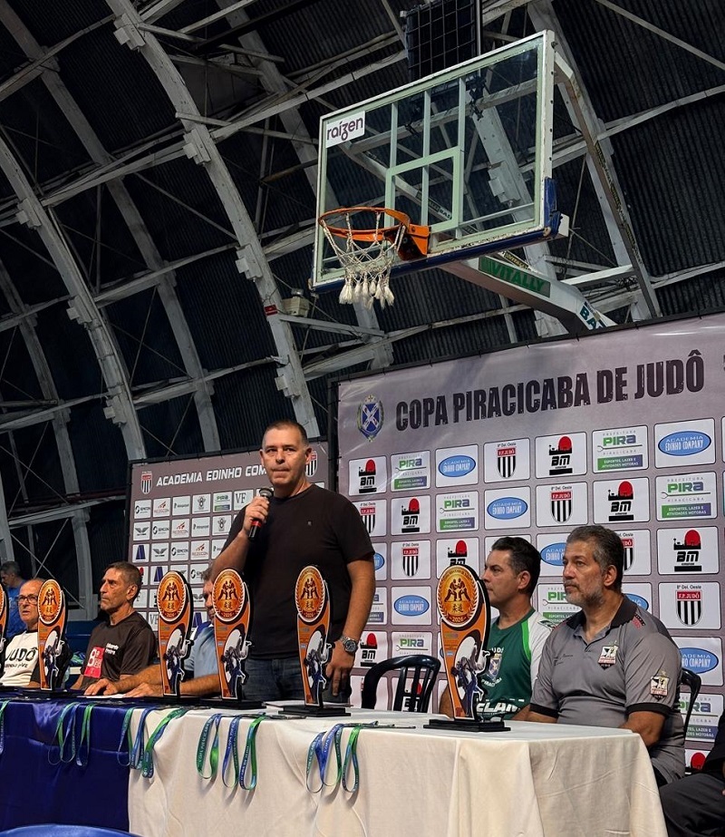 A imagem mostra uma cerimônia ou evento relacionado ao judô, realizada em uma quadra esportiva coberta. No centro da cena, um homem está em pé, segurando um microfone e falando para o público. Ele veste uma camiseta preta. À sua frente, há uma mesa longa coberta por uma toalha branca, sobre a qual estão dispostos vários troféus de diferentes tamanhos e medalhas penduradas, que provavelmente serão entregues aos vencedores da competição. Sentados ao redor da mesa, há outros homens, alguns observando o orador e outros olhando em direção à frente. Atrás deles, uma grande faixa ou banner exibe o nome do evento: "COPA PIRACICABA DE JUDÔ", além de logotipos de patrocinadores e apoiadores, como "ACADEMIA EDINHO COMPANY" e "RAÍZEN". No fundo, é possível ver parte de uma cesta de basquete, indicando que o evento ocorre em uma quadra poliesportiva. A estrutura do teto é metálica e a iluminação é típica de ginásios esportivos. A atmosfera sugere um momento oficial, possivelmente a abertura da competição ou a entrega de premiações.