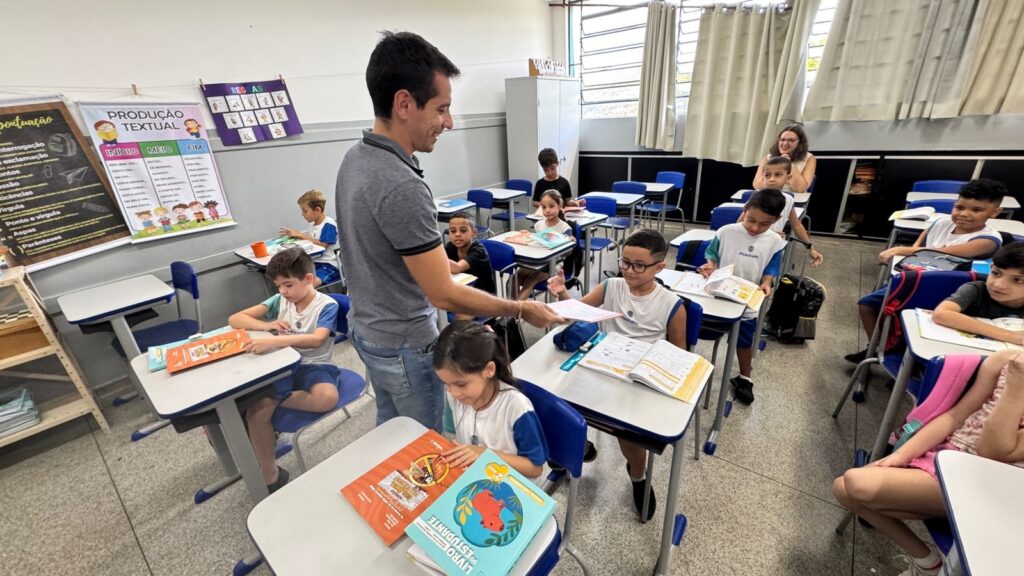A imagem mostra uma sala de aula animada e bem iluminada, provavelmente de uma escola primária no Brasil, dado o uso do português e o estilo do uniforme. No centro da cena, um professor, vestido com uma camisa polo cinza e calça jeans, sorri enquanto entrega um papel a um menino sentado em uma das carteiras. O menino está atento e parece interessado no que recebe. Ao redor, outras crianças, todas vestindo uniformes escolares brancos e azuis, estão sentadas em carteiras azuis e brancas, algumas olhando para o professor, outras para seus livros. A sala é organizada, com paredes brancas e uma faixa cinza na parte inferior. Nas paredes, há cartazes educativos em português; um deles é intitulado "PRODUÇÃO TEXTUAL" e apresenta frases e expressões para ajudar na construção de textos, como "ERA UMA VEZ...", "CERTO DIA...", "DE REPENTE...", entre outras. Outro cartaz indica "REGRAS". Sobre as carteiras, é possível ver livros escolares coloridos, um deles com o título "LIVRO DO ESTUDANTE" e "LÍNGUA PORTUGUESA 2º ANO", indicando que a turma é do segundo ano do ensino fundamental. Ao fundo, há uma mulher, possivelmente uma assistente ou outra professora, observando a aula. A sala tem janelas grandes com cortinas bege, que permitem a entrada de muita luz natural, criando um ambiente acolhedor e propício para o aprendizado. O piso é de azulejo cinza, e o clima geral da imagem é de envolvimento e aprendizado ativo.