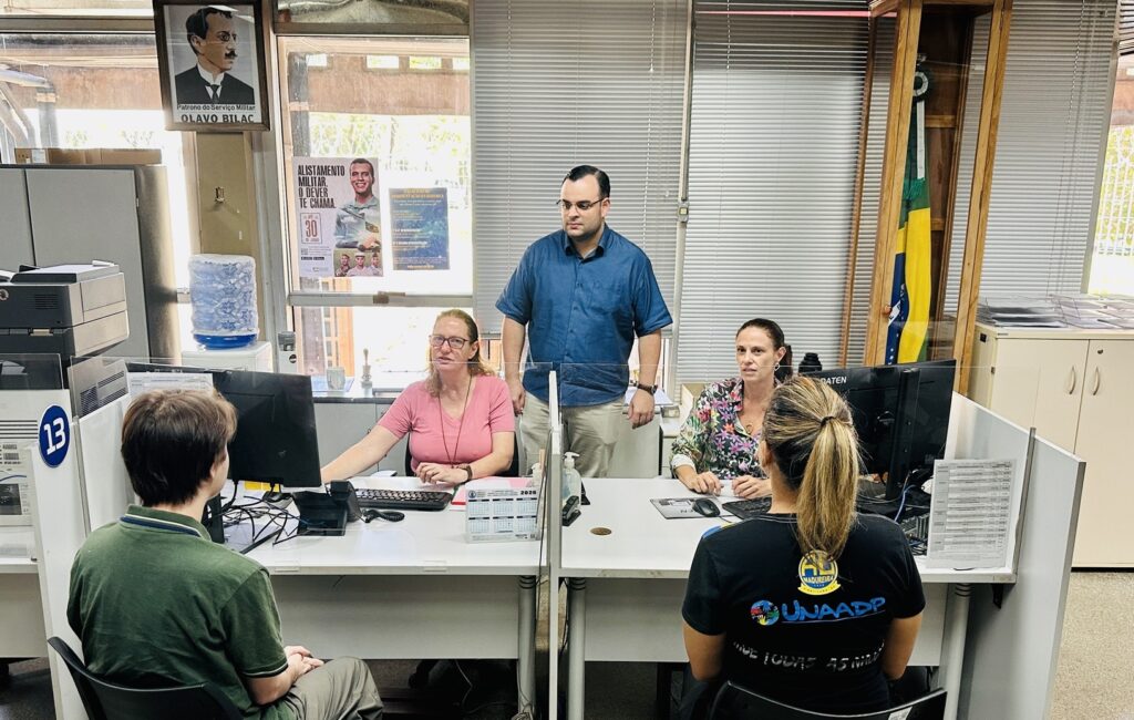 A imagem mostra um ambiente de atendimento público, possivelmente um posto de alistamento militar no Brasil, dado o contexto dos cartazes e símbolos presentes. No centro da cena, há um balcão de atendimento dividido por divisórias de vidro, com duas pessoas sentadas de costas para a câmera: um homem usando uma camisa verde e uma mulher com camiseta preta que tem as inscrições "UNAADP" e "MADUREIRA". Do outro lado do balcão, há três funcionários, dois sentados e um em pé. As duas mulheres sentadas estão trabalhando em computadores, uma veste uma camisa rosa e a outra uma blusa estampada. O homem em pé usa uma camisa azul clara e óculos, e parece estar acompanhando ou auxiliando o atendimento. Ao fundo, há persianas cinzas cobrindo as janelas, uma bandeira do Brasil em um suporte de madeira, e vários cartazes na parede. Um dos cartazes destaca "OLAVO BILAC" como patrono do serviço militar, e outro tem uma chamada para o "ALISTAMENTO MILITAR", com a frase "O DEVER TE CHAMA" e o prazo "ATÉ 30 DE JUNHO". Também há um pequeno calendário de mesa com o ano 2024 visível sobre o balcão. O ambiente é organizado e iluminado, sugerindo um local oficial e voltado para o atendimento ao público, especificamente relacionado ao serviço militar obrigatório.