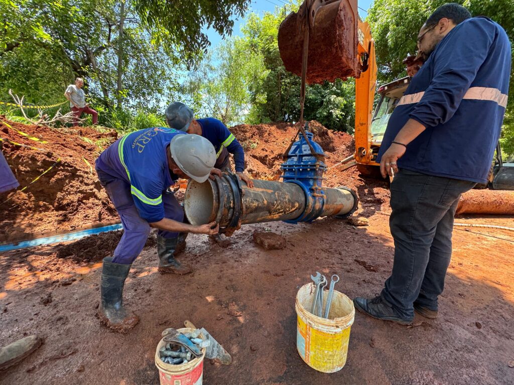 A imagem mostra uma cena de trabalho ao ar livre, durante o dia, em um ambiente de construção ou manutenção de uma tubulação industrial. Três trabalhadores estão no centro da imagem, todos usando capacetes de segurança e roupas de trabalho. Dois deles estão diretamente manuseando uma grande tubulação preta com uma válvula azul, possivelmente realizando instalação ou reparo. Um terceiro homem observa a operação, vestindo camisa azul escura e calça preta. Ao fundo, uma escavadeira de cor laranja está suspensa por uma corrente, segurando a tubulação pesada. O chão está coberto por lama vermelha, indicando que o local pode ser uma obra em terreno úmido ou recentemente escavado. No primeiro plano, há dois baldes plásticos: um contém parafusos grandes e uma chave inglesa, e o outro está cheio de várias chaves inglesas, sugerindo que estão sendo usadas ferramentas manuais para o serviço. Ao fundo, é possível ver uma faixa amarela de segurança e um homem de camiseta cinza e calça vermelha observando a cena. A área está cercada por vegetação verde, com árvores e um céu azul claro, indicando que o trabalho está sendo feito em uma região provavelmente rural ou próxima à natureza. Na roupa de um dos trabalhadores, lê-se "GRP Saneamento Linha Especial", o que sugere que o serviço está relacionado ao setor de saneamento básico, como redes de água ou esgoto. A imagem transmite uma sensação de trabalho pesado, técnica e cuidado na montagem ou manutenção de uma infraestrutura essencial.