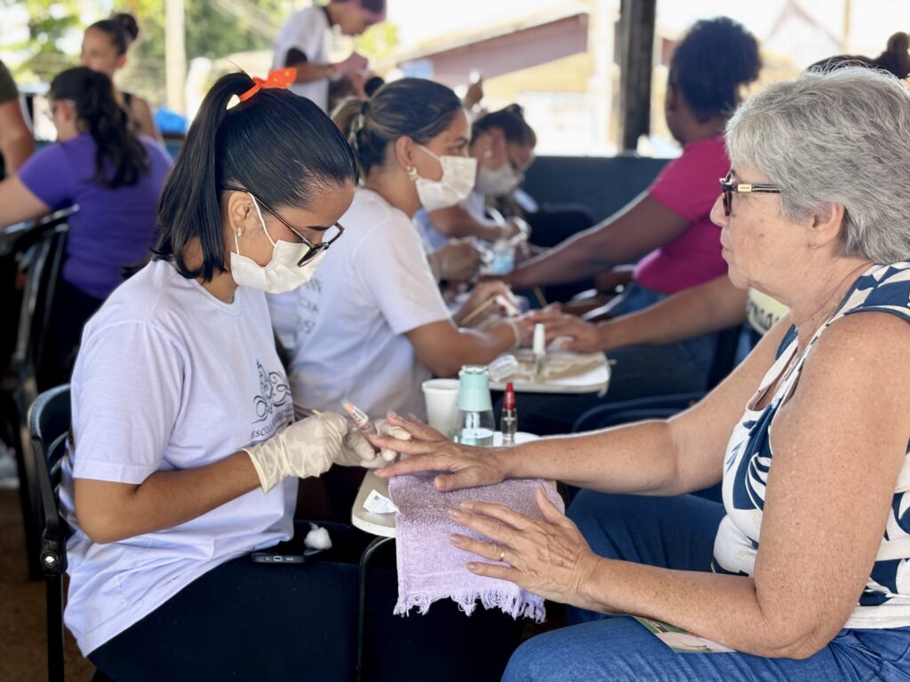 A imagem mostra um ambiente ao ar livre, provavelmente uma ação comunitária ou um evento em uma escola profissionalizante, onde várias mulheres estão recebendo serviços de manicure. Em primeiro plano, há uma jovem com cabelo preso em um rabo de cavalo alto, adornado com um laço laranja. Ela usa óculos, máscara branca e luvas descartáveis brancas, e está aplicando esmalte nas unhas de uma mulher mais velha, que tem cabelo curto e grisalho, usa óculos e uma blusa azul com estampa branca. A mão da mulher mais velha está apoiada sobre uma pequena toalha roxa em uma mesa dobrável. Ao fundo, outras duplas estão realizando a mesma atividade, com as manicures também vestindo camisetas brancas e máscaras, concentradas no trabalho. O ambiente é iluminado pela luz natural do dia, com uma estrutura coberta que protege o local e algumas árvores e edifícios desfocados ao fundo. Na camiseta das manicures, é possível ver parte de um logotipo com a palavra "ESCOLA", indicando que o evento pode estar vinculado a uma instituição de ensino. A foto tem foco seletivo, destacando nitidamente a jovem manicure e a cliente, enquanto o restante do cenário está desfocado. A cena transmite um sentimento de cuidado, aprendizado e interação comunitária.