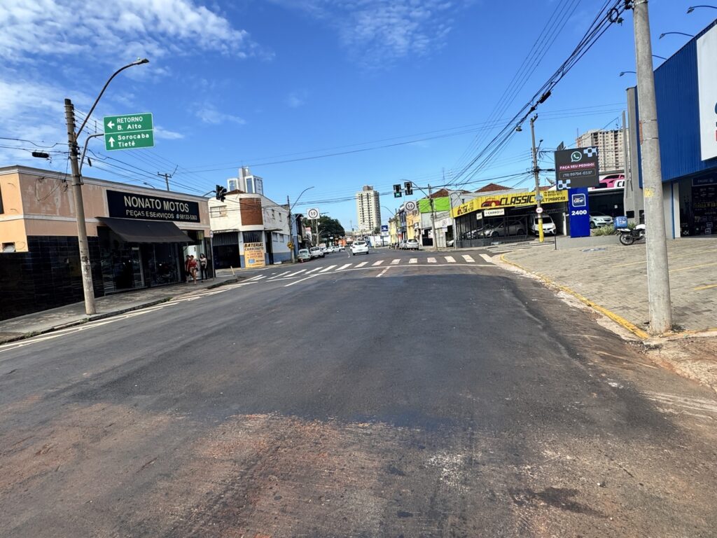 A imagem mostra um cruzamento amplo em uma área urbana, sob um céu azul claro com poucas nuvens. A rua é asfaltada e bastante larga, com várias faixas de tráfego visíveis. À esquerda, há um estabelecimento comercial chamado "NONATO MOTOS PEÇAS E SERVIÇOS", com uma placa que inclui um número de telefone. Ao lado, há outro prédio amarelo com o letreiro "CLASSIC MULTIMARCAS". À direita, destaca-se uma placa verde que indica direções para "RETORNO B. Alto", "Sorocaba" e "Tietê", sugerindo que a foto foi tirada em uma região do estado de São Paulo, Brasil. No fundo, é possível ver prédios mais altos, indicando uma área mais central ou urbana. Também há postes com fiação elétrica e sinais de trânsito. O ambiente aparenta ser movimentado, embora no momento da foto a rua esteja relativamente vazia. O clima é claro e ensolarado, transmitindo uma sensação de dia típico e tranquilo.