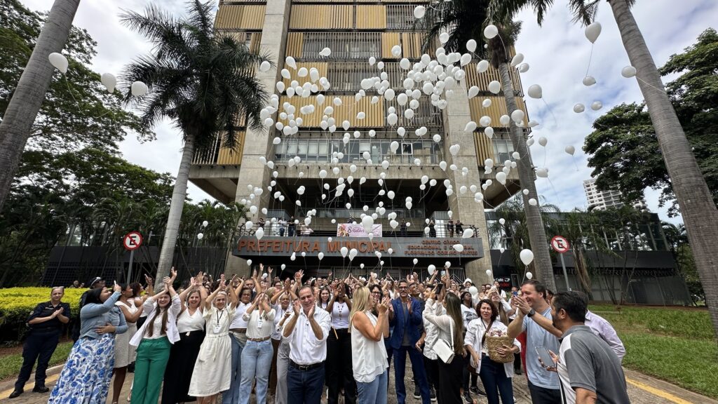 A imagem mostra um grupo grande de pessoas reunidas em frente à fachada de um prédio público identificado como "Prefeitura Municipal". O prédio é moderno, com estrutura em concreto e vidro, e possui uma inscrição em letras grandes e alaranjadas na parte superior que diz "PREFEITURA MUNICIPAL". À direita da entrada principal, há uma placa indicando "CENTRO CÍVICO EDUCACIONAL E CULTURAL FLORIVALDO COELHO PRATES". No centro da imagem, diversas pessoas, a maioria vestindo roupas brancas, estão olhando para cima e soltando balões brancos que estão subindo pelo céu. A expressão das pessoas é de alegria e celebração, algumas aplaudem enquanto outras observam os balões flutuando. À esquerda e à direita, há placas de sinalização de estacionamento proibido. No andar superior do prédio, há um banner com o texto "8 Mulher" e um símbolo rosa, sugerindo que o evento está relacionado ao Dia Internacional da Mulher, comemorado no dia 8 de março. O ambiente é ao ar livre, com algumas palmeiras ao redor e um céu levemente nublado. A foto foi tirada com uma lente grande-angular, dando uma sensação de amplitude e mostrando o prédio imponente sobre as pessoas.