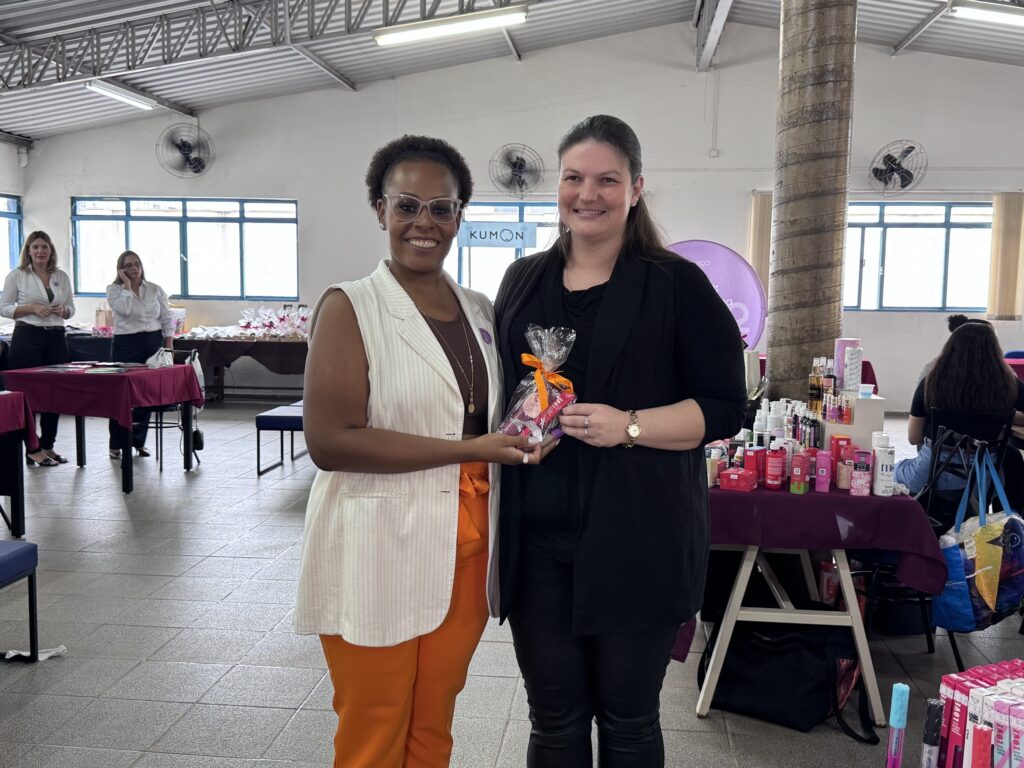 A imagem mostra duas mulheres no centro de um ambiente interno, provavelmente um salão de eventos ou uma escola. Elas estão sorrindo e uma delas está entregando um pequeno presente embrulhado em plástico transparente com uma fita laranja para a outra. A mulher à esquerda usa óculos, uma blusa branca com listras e calças laranja, enquanto a mulher à direita veste roupas pretas e tem o cabelo preso. Ao fundo, há mesas cobertas com toalhas bordô, com diversos produtos expostos, possivelmente cosméticos, e algumas cadeiras. Também é possível ver outras duas mulheres, uma delas falando ao telefone. Na parede, há uma placa azul com o logo "KUMON". O ambiente é iluminado por luz natural que entra pelas janelas. A cena transmite um clima de confraternização ou evento promocional.