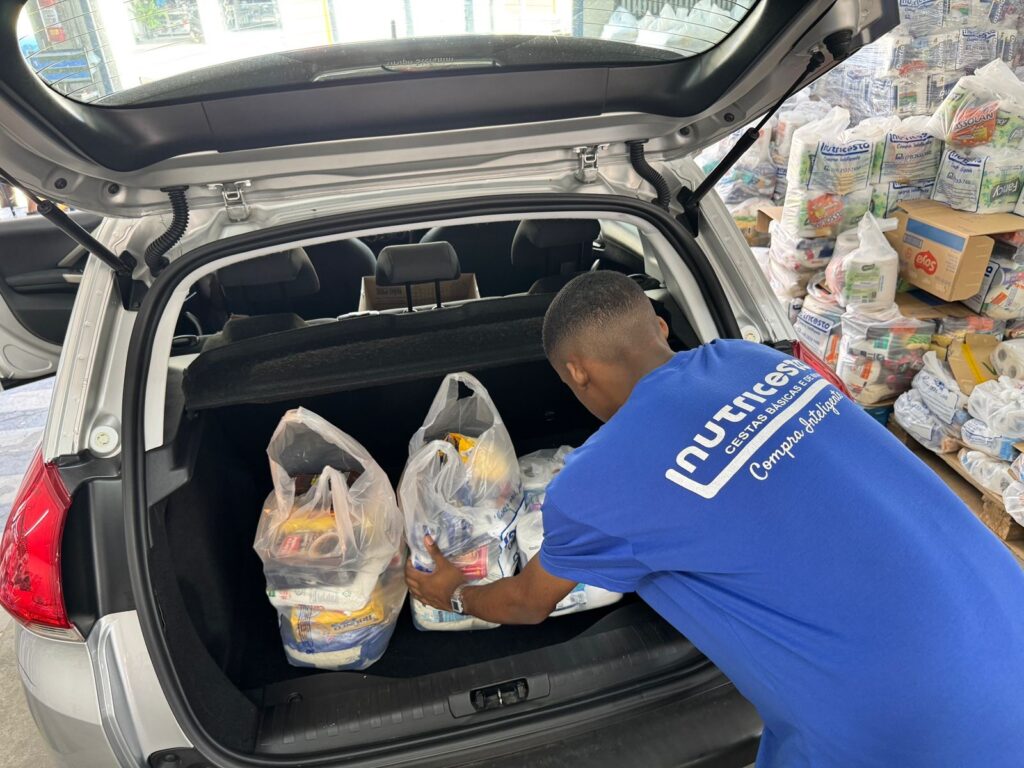 A imagem mostra um homem de camiseta azul carregando sacolas plásticas cheias de mantimentos no porta-malas aberto de um carro prata. Ele está visto de costas e levemente de lado, organizando várias sacolas transparentes com alimentos embalados. Ao lado do carro, há uma grande pilha de cestas básicas embaladas, algumas em plástico transparente e outras em caixas de papelão. No fundo, é possível ver um ambiente que parece ser um depósito ou área de armazenamento, com mais caixas empilhadas. A camiseta do homem tem o logo da empresa "Nutricesta" com o slogan "Compra Inteligente". Os mantimentos nas sacolas incluem itens como café, arroz e outros produtos alimentícios. A iluminação é clara, indicando que a foto foi tirada em um local bem iluminado, possivelmente durante o dia.