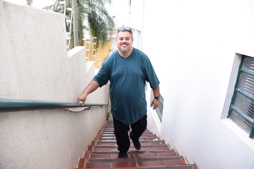 A imagem mostra um homem de meia-idade subindo uma escada externa estreita entre dois prédios. Ele tem cabelos grisalhos, barba por fazer e está sorrindo. Veste uma camiseta azul escura com botões na gola, calças pretas e tênis escuros. Ele usa um relógio no pulso esquerdo e óculos de sol apoiados na cabeça. A escada é feita de azulejos vermelhos com faixas antiderrapantes pretas, e há um corrimão verde escuro à esquerda. Ao fundo, é possível ver uma palmeira e parte de outras construções, indicando que o local é provavelmente uma área urbana em clima quente. A luz é natural e forte, sugerindo que a foto foi tirada durante o dia. A composição utiliza a diagonal da escada para direcionar o olhar para o homem, que está no centro da imagem.