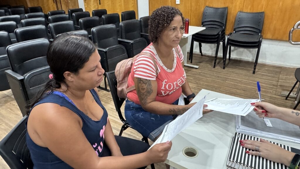 A imagem mostra duas mulheres sentadas à mesa, aparentemente envolvidas em uma conversa ou reunião com uma terceira pessoa, que está parcialmente visível apenas pelas mãos. A mulher à esquerda tem cabelo escuro preso para trás e veste uma regata azul, enquanto a mulher à direita tem cabelo cacheado, veste uma blusa listrada vermelha e branca por cima de uma camiseta cor de rosa com a palavra "forever" escrita, além de shorts jeans. Ambas seguram documentos em mãos, indicando que estão analisando ou discutindo algum material escrito. A mesa está organizada com alguns papéis e um caderno espiralado com capa listrada em preto e branco. Ao fundo, há várias cadeiras pretas alinhadas, sugerindo que o local pode ser uma sala de reuniões, auditório ou espaço para eventos. A iluminação é clara, evidenciando um ambiente interno bem iluminado. A cena transmite uma atmosfera de trabalho, estudo ou atendimento.