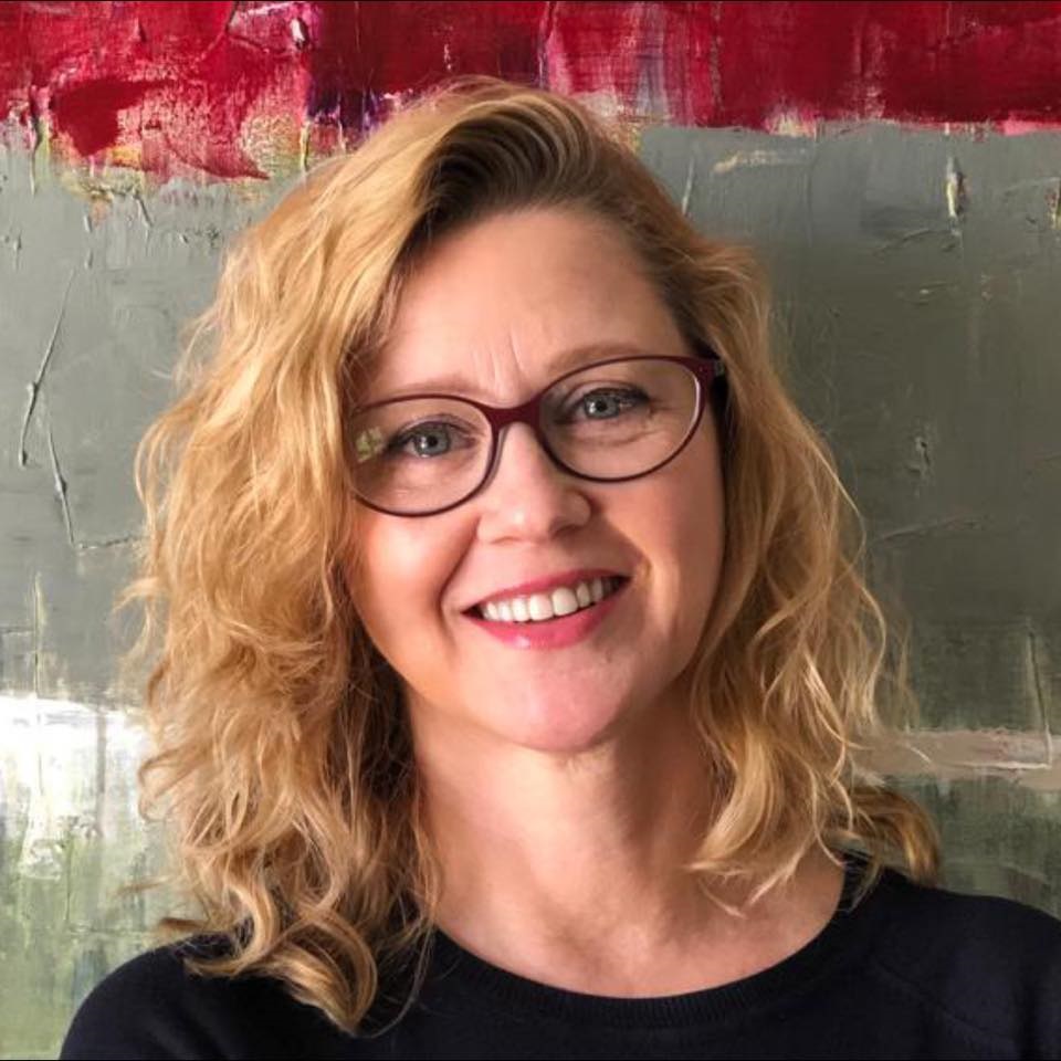 A imagem mostra um retrato próximo de uma mulher de meia-idade, com uma expressão amigável e sorriso suave. Ela tem cabelos loiros ondulados, na altura dos ombros, e usa óculos de armação fina e vermelha. Ela veste uma blusa escura, possivelmente preta ou azul-marinho. O fundo é uma pintura abstrata com tons de verde-oliva, cinza e uma faixa horizontal vibrante em vermelho ou magenta na parte superior. A iluminação é suave, destacando suas feições de forma natural. A composição transmite uma sensação profissional e acolhedora.