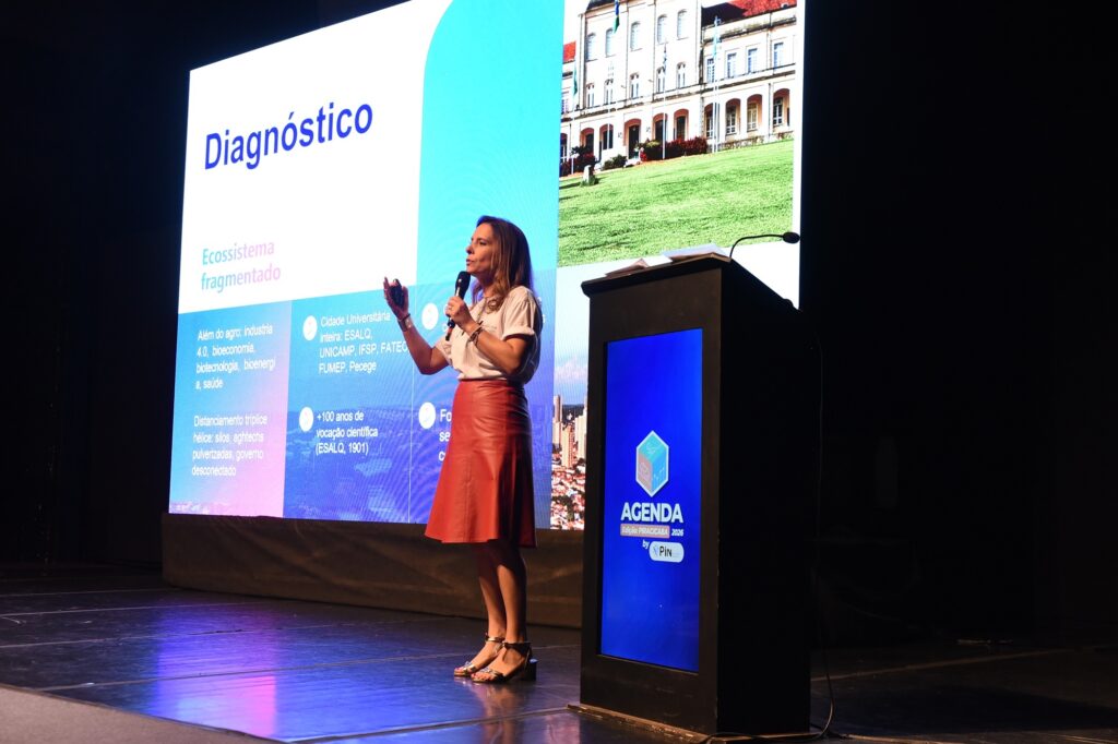 A imagem mostra uma mulher apresentando em um palco durante um evento ou conferência. Ela está vestindo uma camiseta branca e uma saia midi vermelha de couro, além de sandálias douradas de salto. Está segurando um microfone em uma mão e um controle remoto na outra, parecendo explicar ou destacar algum ponto. Ao fundo, há uma grande tela digital que exibe um slide com o título "Diagnóstico" e vários tópicos relacionados ao ecossistema de inovação, mencionando termos como "Ecossistema fragmentado", "Além do agro: indústria 4.0, bioeconomia, biotecnologia, bioenergia, saúde", e "Distanciamento tríplice hélice: silos, agtechs pulverizadas, governo desconectado". Também são citadas instituições acadêmicas como ESALQ, UNICAMP, IFSP, FATEC, FUMEP e Pecege, ressaltando uma vocação científica de mais de 100 anos. Na frente da palestrante, há um púlpito preto com um monitor que mostra a frase "AGENDA Edição: PIRACICABA 2024 by PiN", indicando o nome do evento e sua edição. O ambiente é um auditório escuro, com foco de iluminação na palestrante e na tela, destacando a apresentação. O cenário sugere um contexto profissional e acadêmico, possivelmente relacionado à inovação, ciência e tecnologia no Brasil.