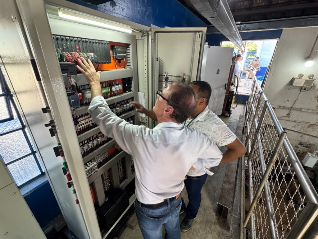A imagem mostra dois homens em um ambiente industrial, provavelmente uma sala técnica ou de manutenção. Eles estão diante de um grande painel elétrico aberto, repleto de fios coloridos e componentes eletrônicos organizados em várias fileiras. O homem mais próximo da câmera, vestido com uma camisa azul clara e jeans, está mexendo no painel, enquanto o outro, com camisa branca estampada, observa atentamente. O espaço é iluminado por uma luz natural que entra por uma janela com grade e também por uma luz artificial interna no painel. Ao fundo, através de uma porta aberta, é possível ver outra pessoa em uma área externa iluminada pelo sol. O ambiente transmite uma sensação de trabalho técnico e inspeção detalhada de equipamentos elétricos.