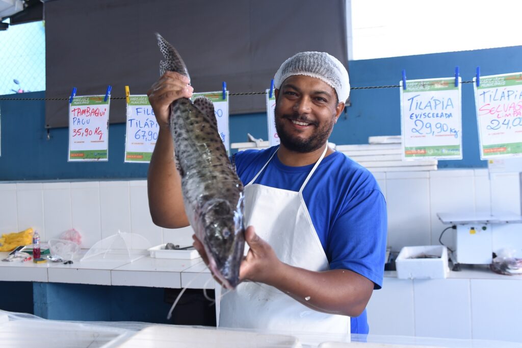 A imagem mostra um peixeiro sorridente segurando verticalmente um peixe grande e manchado, possivelmente um pintado ou surubim, em uma feira ou mercado municipal de peixes. Ele veste uma camiseta azul, avental branco e touca de proteção. Ao fundo, há placas com preços de diferentes tipos de peixes, como tambaqui, pacu e tilápia, indicando valores por quilo em reais. O ambiente parece ser interno, com iluminação natural e azulejos brancos no balcão onde o peixe está apoiado. A atmosfera transmite uma sensação de frescor e confiança, típica de um mercado público de pescados.