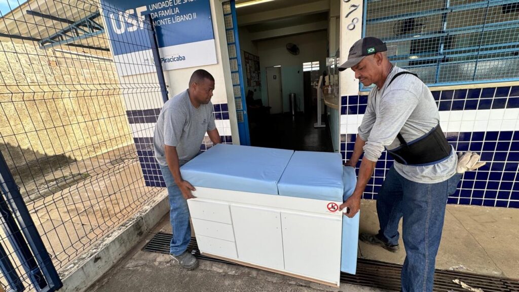 A imagem mostra dois homens carregando um armário médico branco com tampo acolchoado azul-claro para dentro de um prédio. O homem à esquerda veste camiseta cinza e jeans azuis, enquanto o da direita usa camiseta cinza de manga longa, jeans, um cinto de suporte lombar preto e um boné com a bandeira do Brasil. Eles estão na entrada de um prédio identificado por uma placa que diz "USF UNIDADE DE SAÚDE DA FAMÍLIA MONTE LÍBANO II", localizado em Piracicaba. A fachada do prédio é revestida com azulejos azuis e brancos, e há uma grade metálica ao lado esquerdo da entrada. A cena é iluminada pela luz natural do dia, mostrando o esforço físico envolvido na entrega do equipamento médico a um centro de saúde comunitário.
