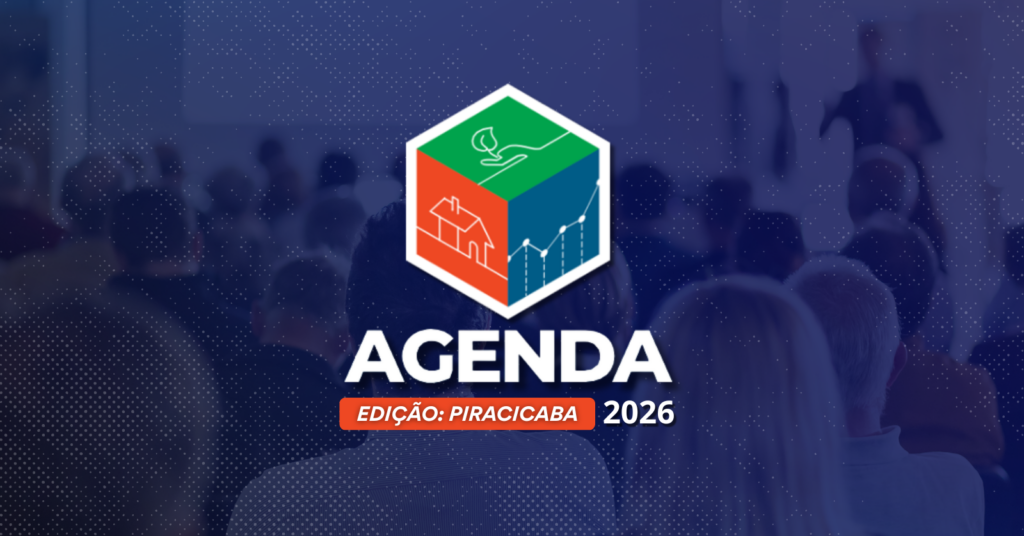 A imagem apresenta um logotipo colorido em forma de cubo dividido em três partes: na seção verde, há uma mão segurando uma folha, simbolizando sustentabilidade; na vermelha, aparece o contorno de uma casa, remetendo a urbanismo ou habitação; e na azul, um gráfico de linhas, sugerindo desenvolvimento econômico ou progresso. Ao fundo, vê-se uma plateia desfocada assistindo a uma apresentação ou conferência, transmitindo o ambiente de um evento formal. Logo abaixo do cubo, está escrito em destaque “AGENDA” e, em seguida, “EDIÇÃO: PIRACICABA 2026”, indicando tratar-se de uma iniciativa ou encontro voltado para o planejamento e futuro da cidade.