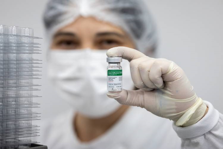 A imagem mostra um profissional de saúde ou laboratório segurando um pequeno frasco de vacina em direção à câmera. O frasco é de vidro com uma tampa prateada e possui um rótulo verde e branco, onde se lê "Influenza" e "10 doses". O profissional está usando máscara facial branca, touca e luvas, todos brancos, em um ambiente que parece ser um laboratório ou uma instalação de fabricação farmacêutica, com fundo branco e iluminação clara. Ao lado esquerdo da imagem, há uma pilha de bandejas plásticas transparentes. A imagem utiliza um foco seletivo, deixando o frasco em destaque nítido, enquanto o rosto do profissional e o fundo estão desfocados. O cenário transmite um ambiente clínico e seguro, relacionado à produção ou distribuição de vacinas.